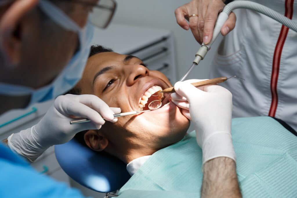 Dentist examining oral cavity of young African American man working in in dental clinic with assistant.